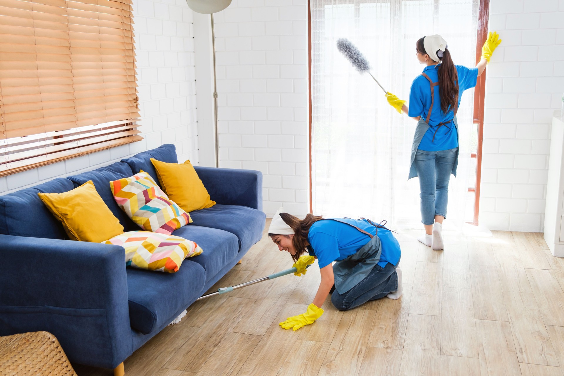 Young professional cleaning service women workers working in the house. Girls housekeeper wiped a wet mop under the sofa and another one cleans the window. Cleaner and chores concept